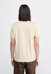 Short-sleeve, crew neck t-shirt in beige cotton, smooth texture, curved hem, worn with brown pants, viewed from the back.