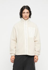 Veste en polaire couleur crème clair avec un col haut, fermeture éclair à l'avant et une surface texturée. Elle comprend une poche poitrine avec une fermeture éclair blanche.