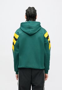 Osoba s vrkočmi, oblečená v zelenej mikine s žltými a čiernymi pruhmi na rukávoch, stojaca chrbtom k jednoduchému pozadiu.