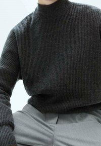 Personne portant un pull en maille côtelée gris foncé avec col montant et un pantalon plissé gris clair, assise devant un fond uni.
