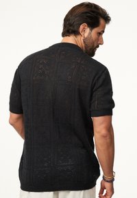 Schwarzes, gestricktes Oberteil mit kurzen Ärmeln, das ein strukturiertes, offenes Muster mit quadratischen Motiven und dezenten Blumenmustern aufweist. Rundhalsausschnitt und lässige Passform.