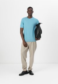 Svetlo moder pulover s kratkimi rokavi z rebrasto teksturo, v kombinaciji z bež nabranimi hlačami in črnimi usnjenimi čevlji, drži črno jakno.