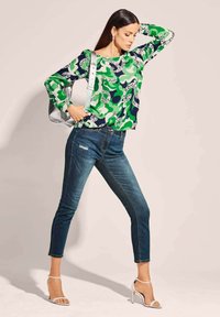 Grünes, gemustertes Langarmoberteil mit abstrakten Formen, kombiniert mit eng anliegenden blauen Jeans mit einem destroyed Detail und weißen, geschnürten High Heels.