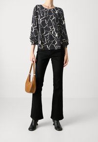 Personne portant un haut à motifs abstraits noir et blanc, un pantalon flare noir, des bottines noires, tenant un sac à main marron par les poignées.