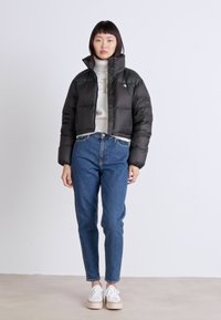 Svart pufferjakke med kort design, glidelås, og høy krage, båret over en kremfarget genser og blå jeans, kombinert med hvite sko.