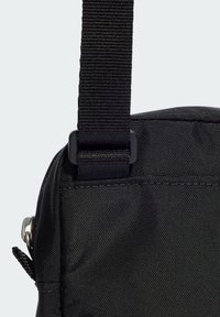 Svart väska med en texturerad nylonutsida, som har ett hållbart bärrem med plastspänne och dragkedja. Detaljerad sömnad längs kanterna.