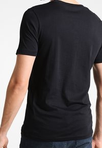 Osoba v jednobarevném černém tričku s krátkým rukávem zobrazená zezadu, stojící na bílém pozadí.