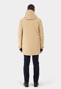 Beige gewatteerde jas met een rits, voorzien van een iets gewatteerde textuur en verstelbare manchetten, gecombineerd met donkere denim broek en zwarte schoenen.