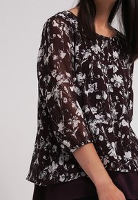 Durchsichtige lila Bluse mit weißem Blumenmuster, runder Halslinie und Dreiviertelärmeln; weiche Textur, fließendes Design.