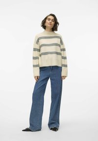 Maglione crema con strisce orizzontali grigie, maniche larghe e colletto alto. Abbinato a jeans in denim blu a vestibilità ampia e scarpe nere.