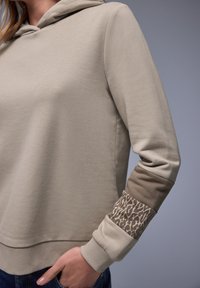 Femme portant un sweat à capuche beige avec un imprimé léopard et des panneaux de manches de couleur unie, main dans la poche, devant un fond gris uni.
