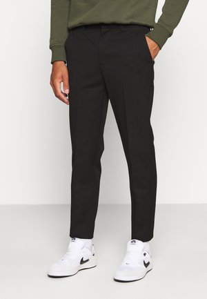 Mann trägt schwarze Slim-Fit-Hose, weiße Nike-Sneaker und ein olivgrünes Langarmshirt, steht vor einem einfarbigen Hintergrund.