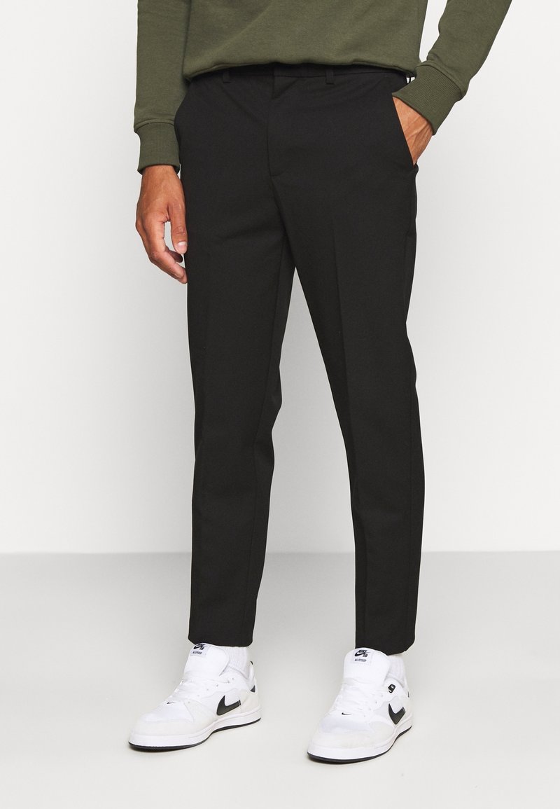 Homme portant un pantalon noir slim-fit, des baskets Nike blanches et un haut à manches longues vert olive, debout devant un fond uni.
