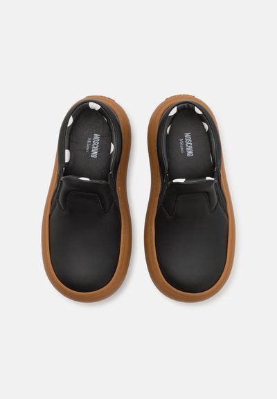Melni slip-on apavi ar gludu ādas virsu un noapaļotu brūnu gumijas zoli. Iekšpusē ir balto punktiņu raksts. Redzama zīmola identifikācija.