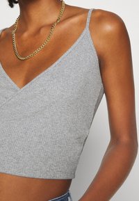 Top court en maille côtelée grise avec un décolleté en V et des bretelles fines, associé à un collier chaîne dorée, mettant en valeur le tissu texturé et la coupe ajustée.