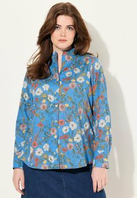 Camisa azul con botones y mangas largas, con un patrón floral en rojo, blanco, amarillo y verde, hecha de una tela ligera.