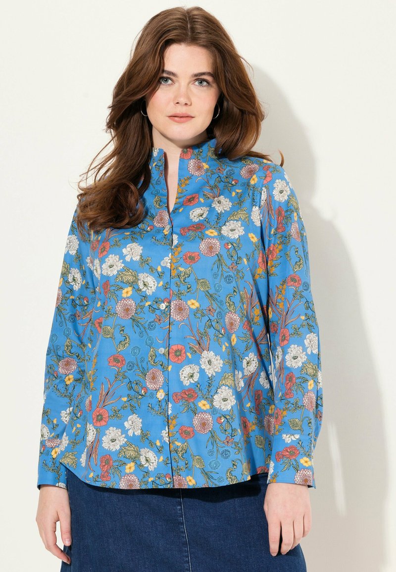 Camisa azul con botones y mangas largas, con un patrón floral en rojo, blanco, amarillo y verde, hecha de una tela ligera.