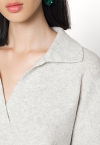 Vrouw in een zacht lichtgrijze gebreide top met een brede kraag en diepe V-hals, met donker haar en groene kralenoorbellen zichtbaar.