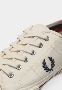 Gros plan sur une basket en toile blanche avec des lacets blancs, des œillets argentés et un logo de couronne de laurier brodé en noir sur le côté.