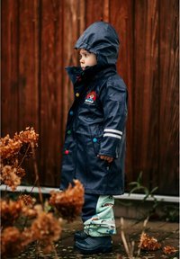 Laineen vedenpitävä sadetakki, jossa on moniväriset napit, huppu ja valkoiset raidat. Yhdistetty vaaleanvihreisiin housuihin, joissa on eläinkuvioita ja mustiin saappaisiin.