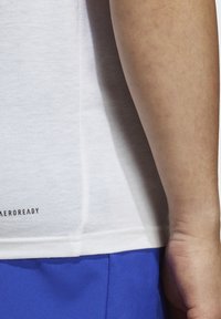 Hvid atletisk t-shirt lavet af letvægtsstof, med en subtil tekstur og trykt "AEROREADY" label i bunden. Blå shorts synlige nedenunder.