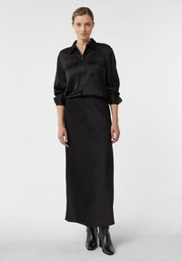 Kobieta w czarnej koszuli zapinanej na guziki, włożonej w długą czarną spódnicę, w połączeniu z czarnymi botkami za kostkę, stojąca na jednolitym tle.