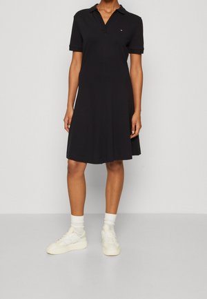 Kobieta ubrana w czarną sukienkę polo na krótkim rękawie, białe skarpetki i białe trampki, stojąca na tle jednolitego jasnoszarego tła.