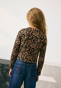 Pikkade blonde juustega laps, kes kannab pruun-mustade leopardimustriga pikkade varrukatega pluusi ja siniseid teksaseid punase südame tikandiga tagataskus.