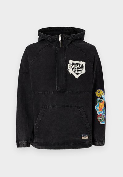 Melnas denima džemperis ar kapuci, ar centrālo rāvējslēdzēju, kenguru kabatu un krāsainiem auduma plankumiem labajā piedurknē.