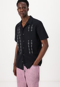 Svart t-skjorte med korte ermer og hvit sikksakk-broderi, med brystlomme og brune knapper; parret med lyserøde jeans.