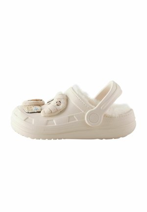 Light beige clog with fluffy inner lining, adjustable heel strap, and decorative plush and plastic charms on the upper.