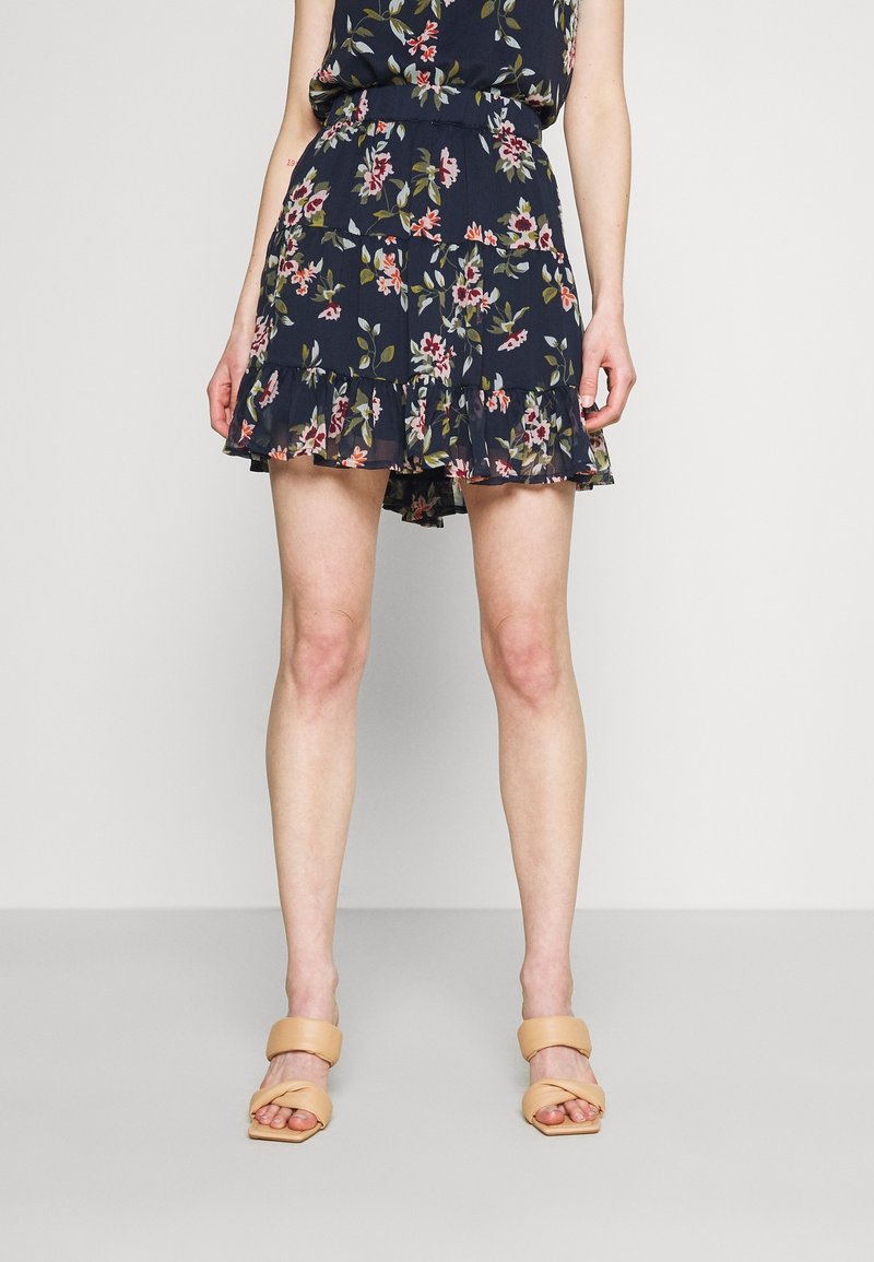 Jupe imprimée florale avec un fond bleu marine foncé, ourlet à volants et taille élastique. Associée à des sandales à talons blocs nude.