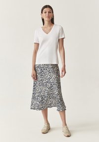 T-shirt blanc à manches courtes en V associé à une jupe bleu marine avec un motif de feuilles en crème. Le mannequin porte des baskets beiges. Design minimaliste.