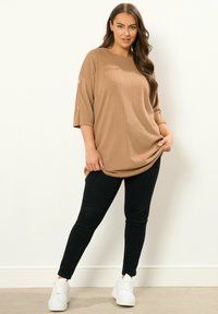 Beige, geripptes Oversized-Oberteil mit Dreiviertelärmeln und Knopfakzenten, kombiniert mit schwarzen Slim-Fit-Hosen und weißen Sneakers.