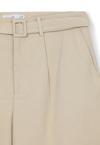 Pantalons beiges avec un devant plat, des passants de ceinture et une ceinture assortie. Fabriqués dans un tissu lisse avec une coupe ajustée et deux poches latérales.