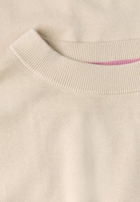 Gros plan d'un col de pull beige tricoté avec des bordures côtelées et une fine ligne rose à l'intérieur de l'encolure.