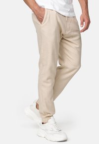 Beige joggerbyxor i lätt tyg, med sidofickor och avsmalnande ben. Kombinerat med vita sneakers mot en neutral bakgrund.