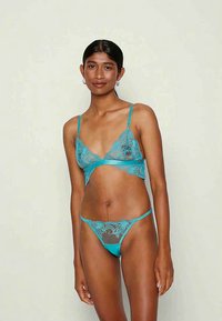 Mulher de pé contra um fundo simples, vestindo um conjunto de lingerie em renda teal com bordados florais, olhando para a câmera com um leve sorriso.