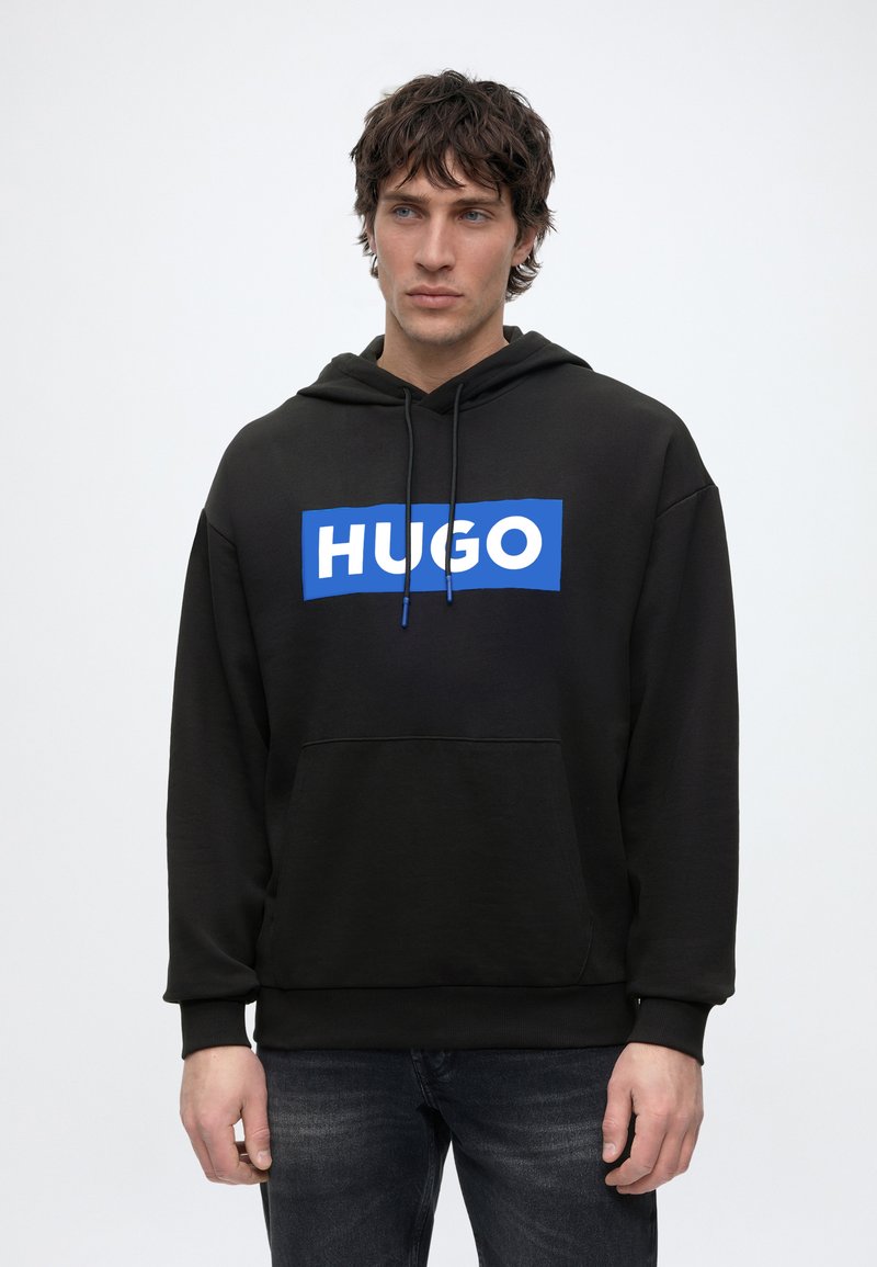 Junger Mann trägt schwarzen Hoodie mit blau-weißem "HUGO"-Logo, steht vor einfachem hellem Hintergrund und schaut leicht nach rechts.