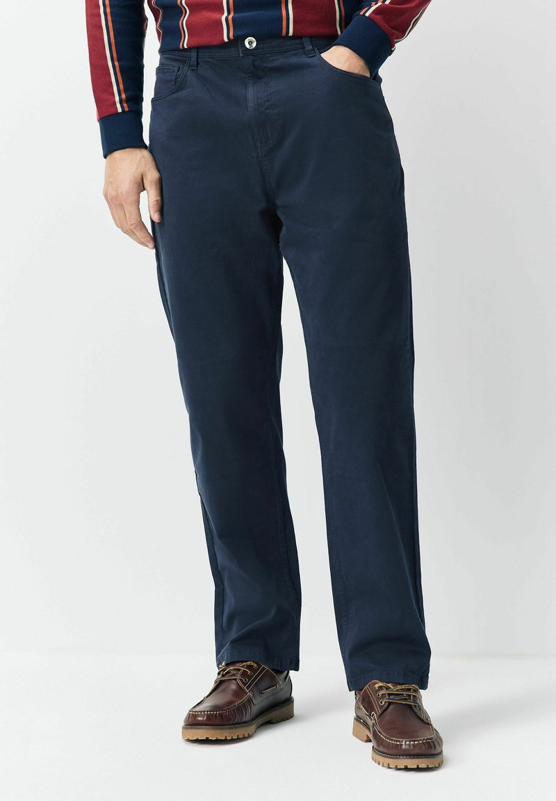 Mann trägt eine marineblaue Hose und braune Lederschuhe und steht mit einer Hand in der Tasche vor einem schlichten weißen Hintergrund.