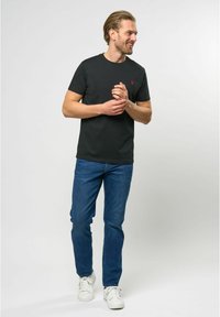 Juodas medvilninis marškinėliai su apvaliu kaklu, su mažuoju raudonu logotipu. Derinama su mėlynomis džinsinėmis kelnėmis ir baltais sportbačiais, stovintis prieš neutralaus fono.