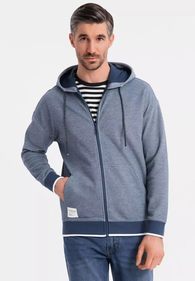Haut à capuche zippé pour homme en bleu foncé avec une capuche, des poches kangourou et des accents contrastants en marine. Fabriqué en tissu doux et texturé. Chemise rayée en dessous.