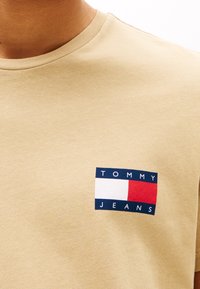Camiseta de algodón beige con un parche rectangular en azul marino, rojo y blanco que lleva el texto "TOMMY JEANS" en la parte izquierda del pecho.