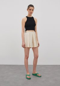 Zwarte crop top met zijstrikjes, crèmekleurige geplooide minirok en groene slippers. Gladde texturen met een aansluitend ontwerp.