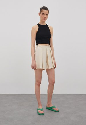 Schwarzes Crop-Top mit seitlichen Bindebändern, cremefarbene, plissierte Mini-Rock und grüne Flip-Flops. Glatte Texturen mit einem figurbetonten Design.