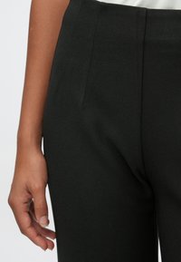 Close-up da mão de uma pessoa descansando ao seu lado, vestindo calças pretas justas e uma blusa branca.