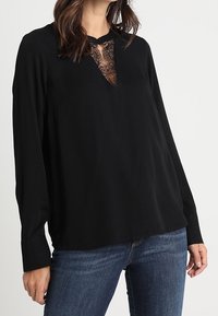 Blusa nera a maniche lunghe con pannello in pizzo all'collo, vestibilità rilassata e texture di tessuto liscia. Abbinata a jeans blu.