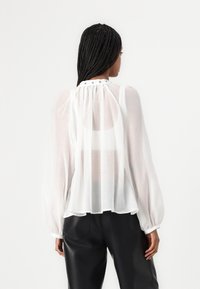 Blusa bianca trasparente con maniche lunghe, dettagli arricciati nel collo e una silhouette morbida. Indossata con pantaloni in pelle nera.