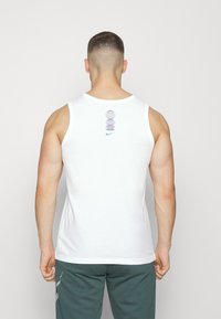 Man met kort haar, gekleed in een witte mouwloze tanktop met een blauw Nike-logo en een geometrisch ontwerp op de bovenrug, gecombineerd met donkergroene broek.