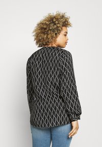 Schwarze Bluse mit langen Ärmeln und weißem ovalem Muster, aus weichem Stoff und mit gerafften Ärmeln. Trägt sie mit hellblauen Jeans, von hinten betrachtet.